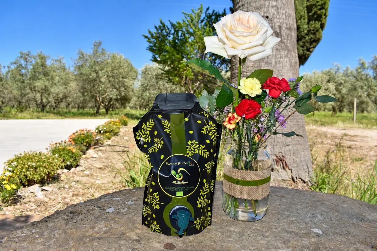Il pouch-up dell'olio RoccaforteBio fra i fiori, con l'uliveto dietro